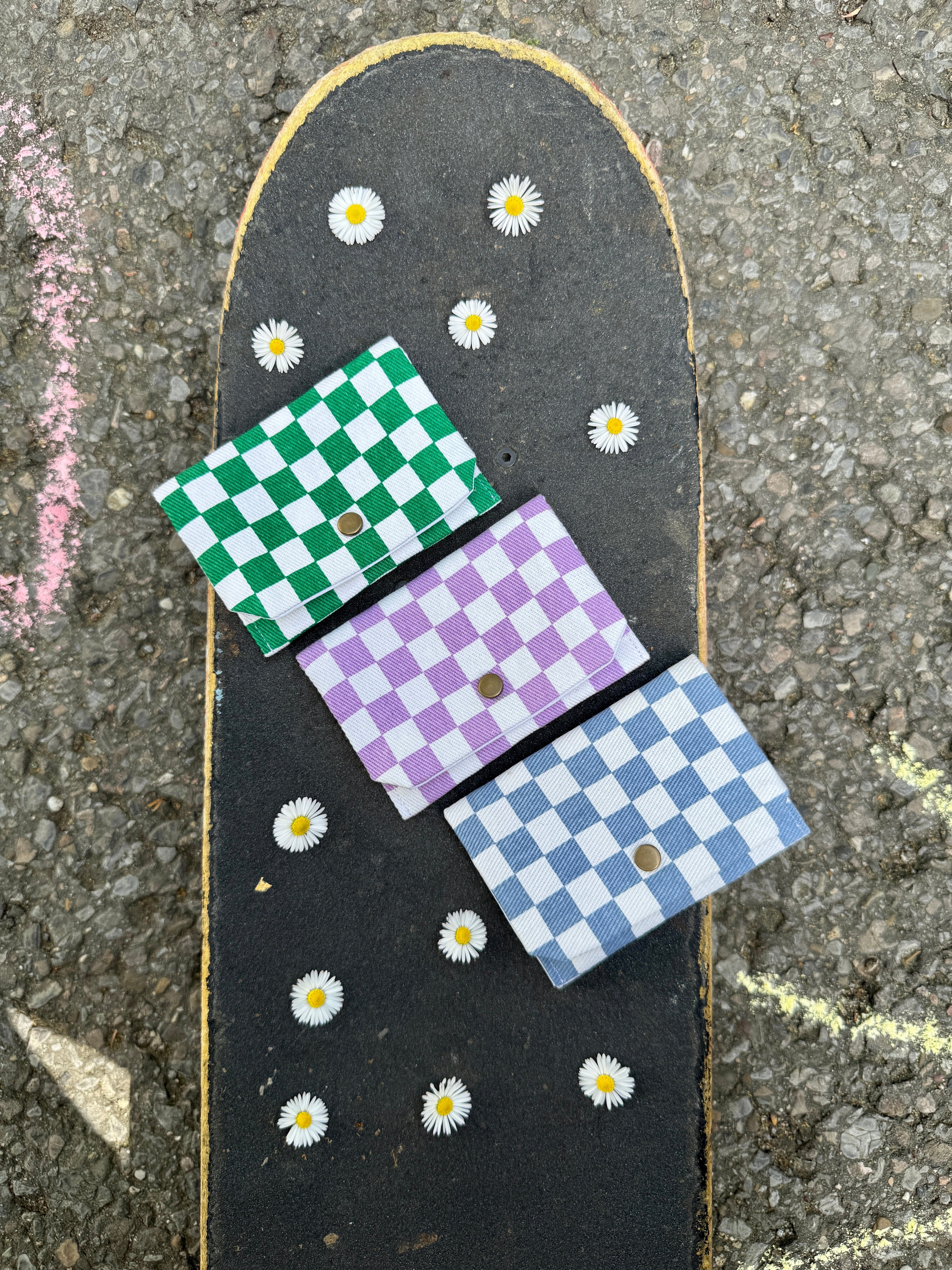 Drei Geldbeutel liegen auf einem Skateboard, umrandet von Gändeblümchen. Die Geldbeutel haben alle ein Checkermuster. Ein Geldbeutel ist grün, ein Geldbeutel lila und ein Geldbeutel blau.