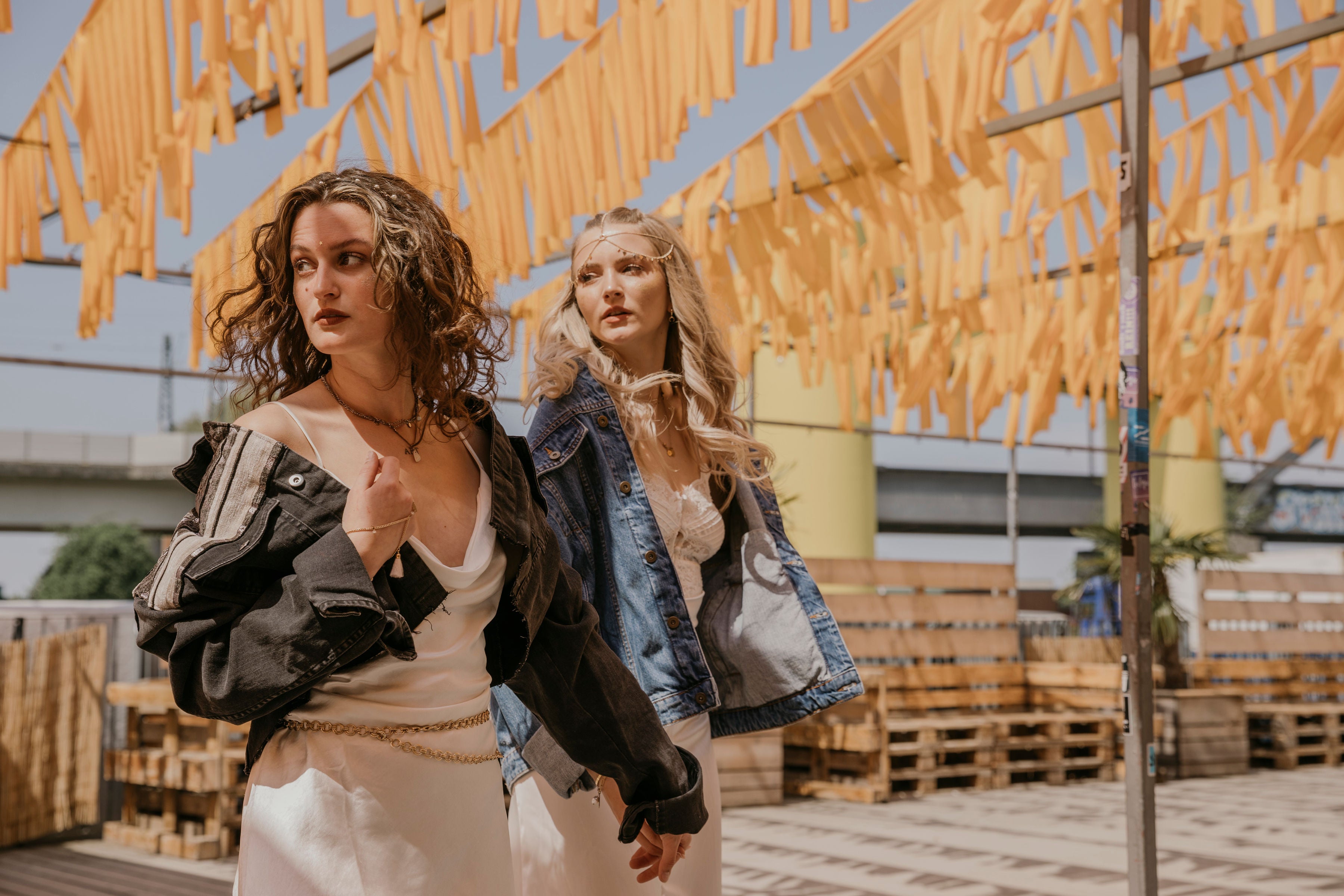 Ein blondes und ein Brunettes Mädchen laufen Hand in Hand über ein Festivalgelänge und schauen sich um. Dabei tragen sie Festival-Outfits von Moonside Collection. Es sind Vintage Jeansjacken, die durch Upcycling zu Einzelstücken gemacht wurden.