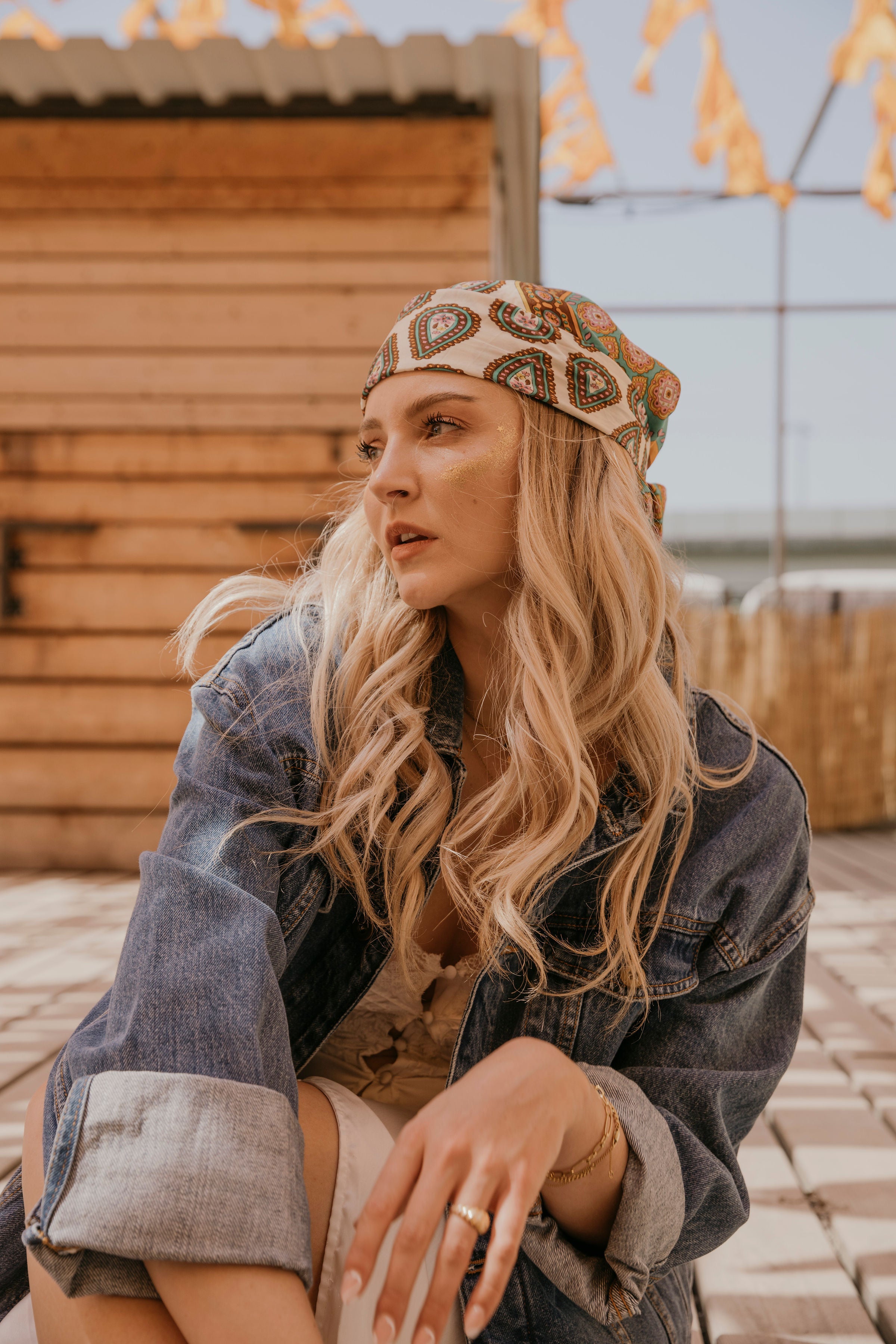 Blondes Mädchen im Hippie-Look sitzt auf dem Boden eines Festival-Geländes. Schaut in die Kamera.