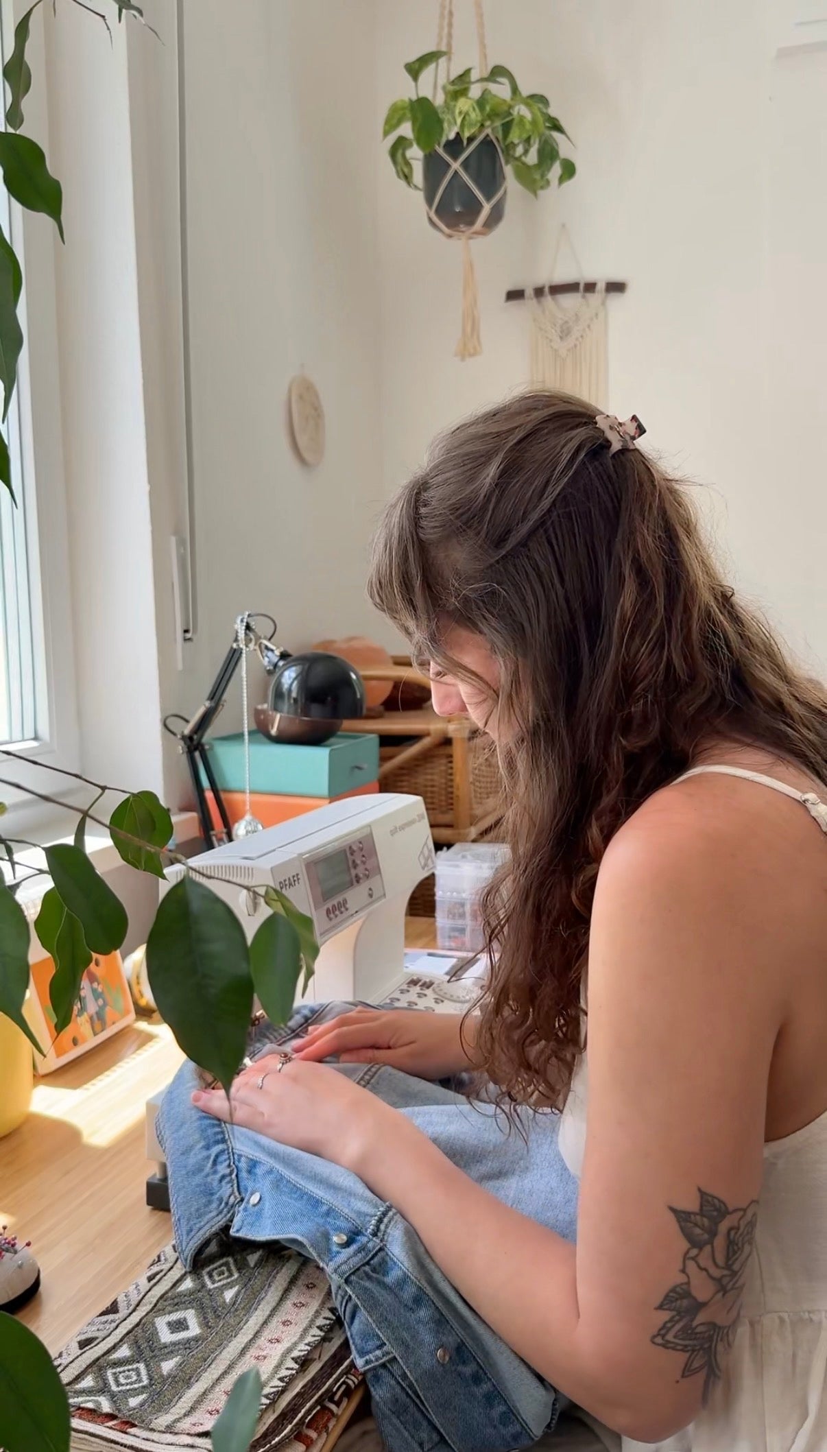 Bild von der Künstlerin Hannah Natterer an ihrem Arbeitsplatz. Sie sitzt an der Nähmaschine und bearbeitet einen Boho-Stoff. Sie ist umgeben von Pflanzen.