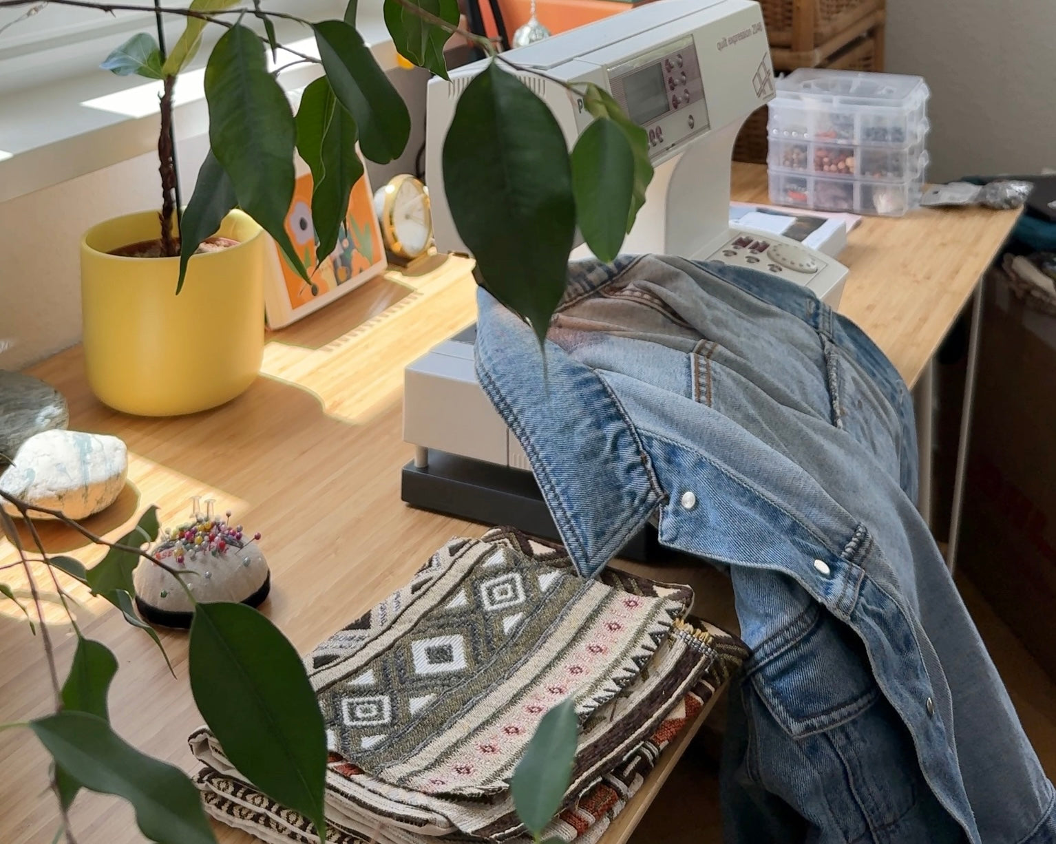 Ein Bild vom Arbeitsplatz von Moonside Collection. Eine Nähmaschine steht auf dem Tisch, ein Stoff mit Boho-Muster liegt in der Maschine. Es sind außerdem Pflanzen und eine Lampe auf dem Tisch.